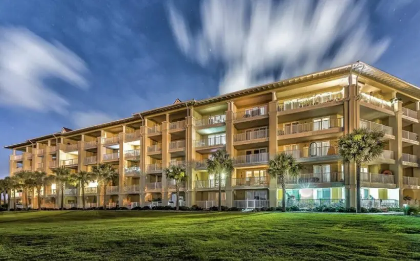 Exterior view of the backside of a five-story apartment building with balconies overlooking a wide lawn and palm trees.