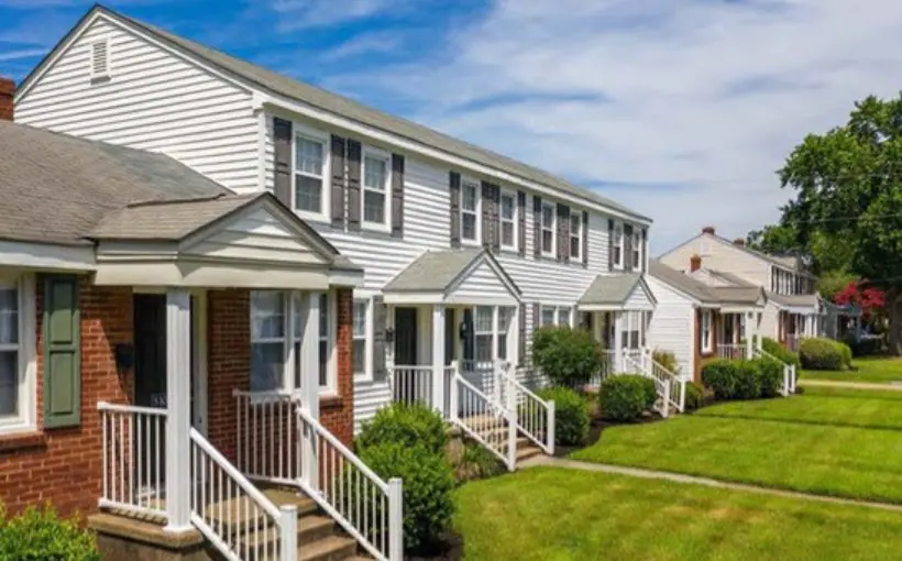 Exterior image of a row of brick and wood landscaped townhomes with stairs leading to covered entrance landings and facing a green lawn