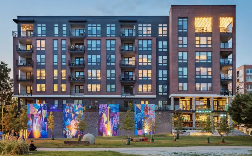 Exterior view of brick mid-rise apartment building facing a reflecting area with seating and urban artwork on vertical concrete panels