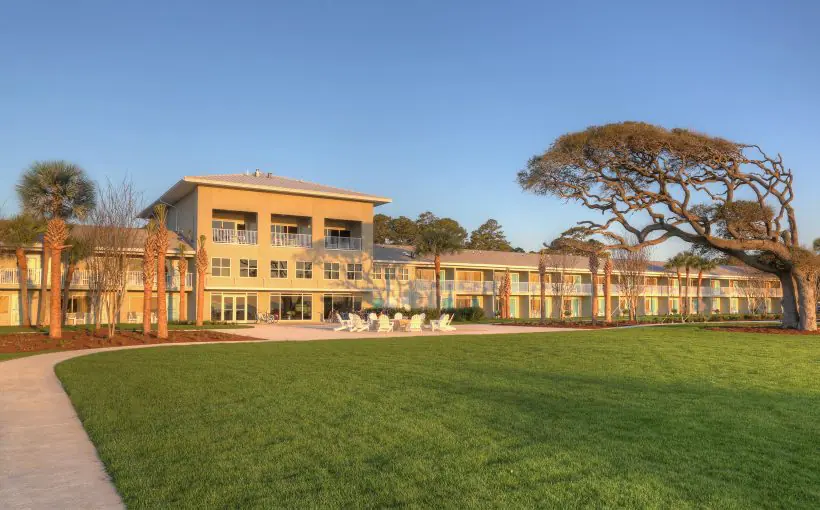 Exterior view of two- and thee-story hotel overlooking seating area and lawn anchored by large ocean wind-swept tree