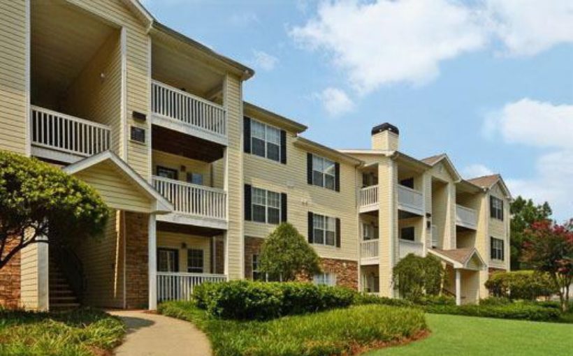 Front exterior view of stone and paneling three story apartment building with shutters, terraces, and manicured landscaping