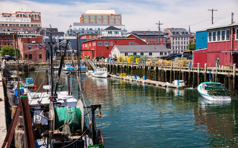 Docked fishing boats in front of colorful waterfront and progressively taller buildings in the background, leading to high rises in the far distance