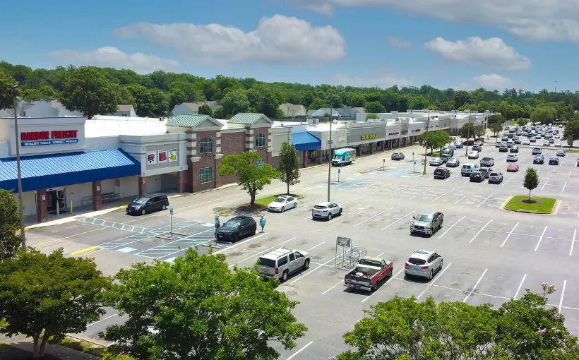 Long range exterior view of a long single-story building containing many shops, along with trees behind and parking in front